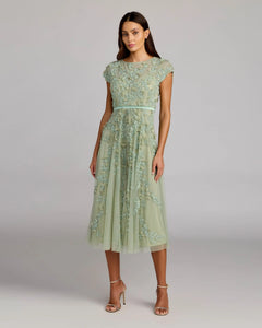 A woman wears the Mac Duggal 94339 midi wedding guest dress in sage, featuring cap sleeves, floral embroidery, a thin belt, and strappy heels—an elegant choice for a wedding reception.