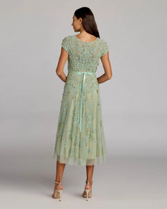 A woman stands in heels, facing away from the camera, wearing the Mac Duggal 94339 Midi Wedding Guest Dress—a light green a-line midi with short sleeves and a ribbon-tied waist—against a plain background.