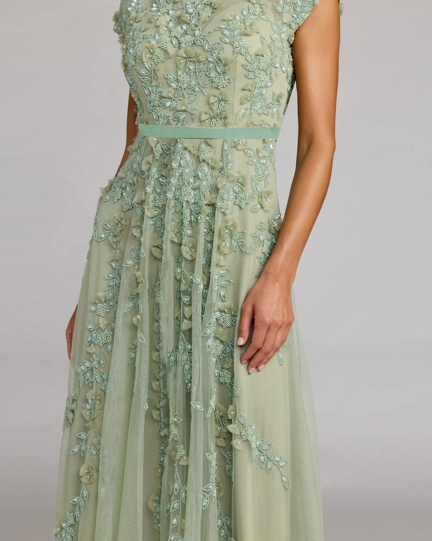 A woman models the Mac Duggal 94339 midi wedding guest dress, featuring light green cap sleeves, floral embroidery, embellishments, and a fitted waist, standing before a plain background.