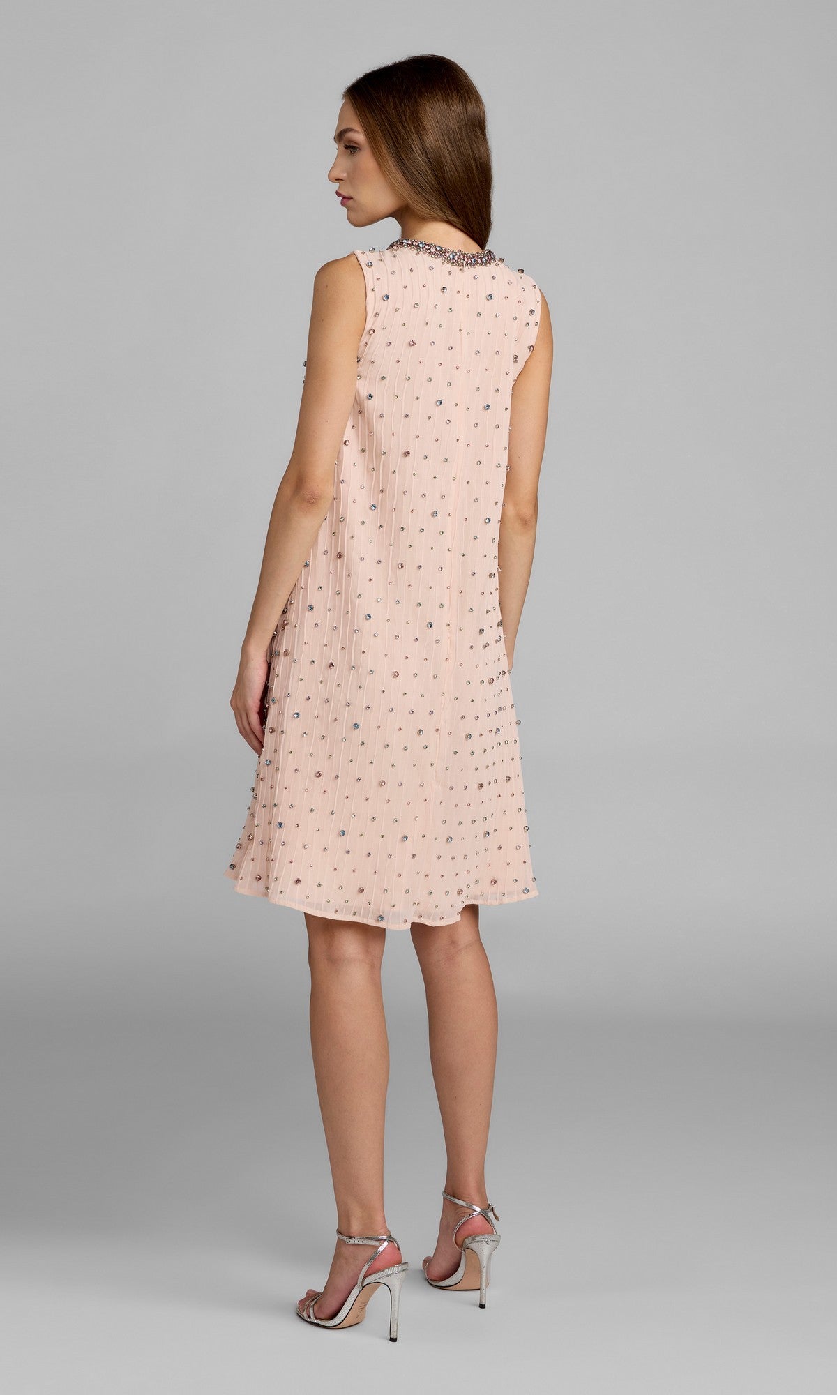 A woman stands with her back to the camera, modeling the knee-length shift party dress Mac Duggal 94354—a sleeveless, light pink design adorned with small gem embellishments—paired with silver high-heeled sandals against a plain gray background.