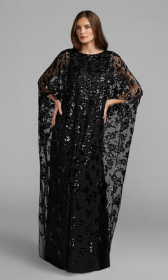 A woman wearing the Mac Duggal 9441 Cape-Sleeve Long Formal Dress in black, featuring wide batwing sleeves and a sheer, sequin-embellished overlay, stands against a plain gray background.