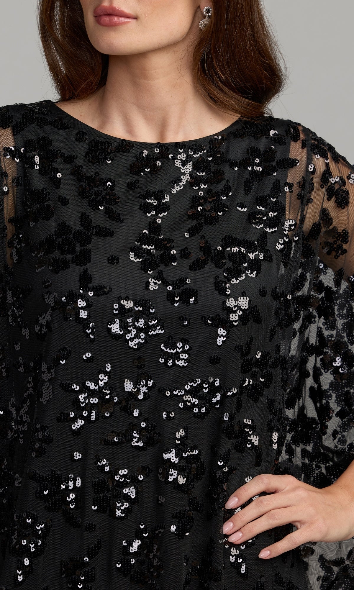 A woman models the Mac Duggal 9441 Cape-Sleeve Long Formal Dress, featuring black sheer cape sleeves and sparkling black and silver sequins, with one hand on her hip; shown from shoulders to mid-thigh.