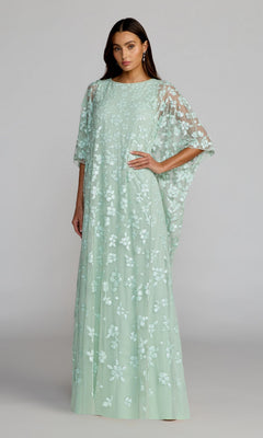 A woman wearing the Mac Duggal 9441 long formal dress in seafoam—a floor-length gown featuring floral sequins, a sheer overlay, and loose cape sleeves—stands against a plain gray background.