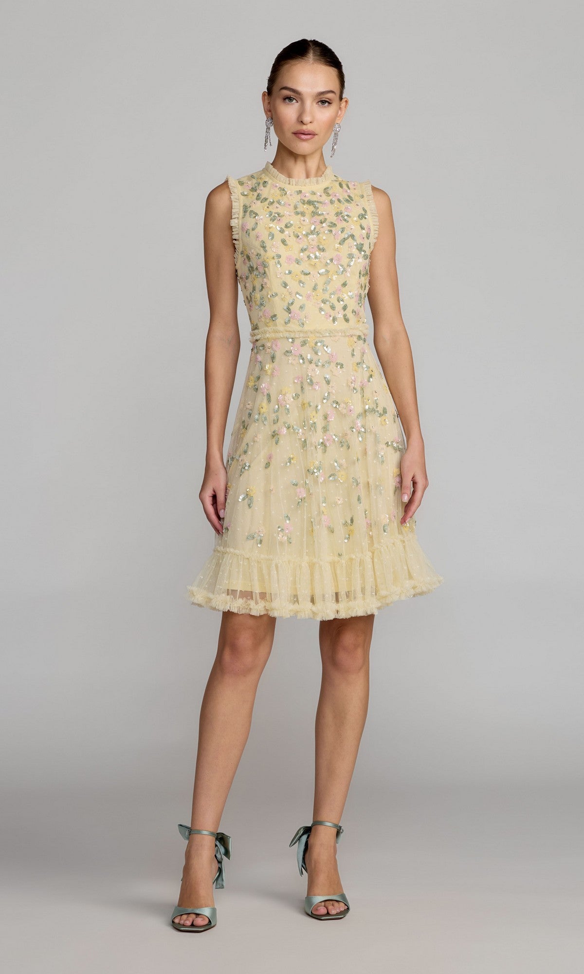 A woman wears the Mac Duggal 9474 Knee-Length Floral-Sequin Dress in lemon, featuring a sleeveless A-line silhouette, floral embroidery, ruffled hem, and paired with green high-heeled sandals.