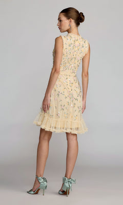 A woman stands in profile wearing the Mac Duggal 9474 Knee-Length Floral-Sequin Dress, featuring a lemon yellow hue, floral embroidery, ruffled hem, and paired with mint green heels tied with bows.