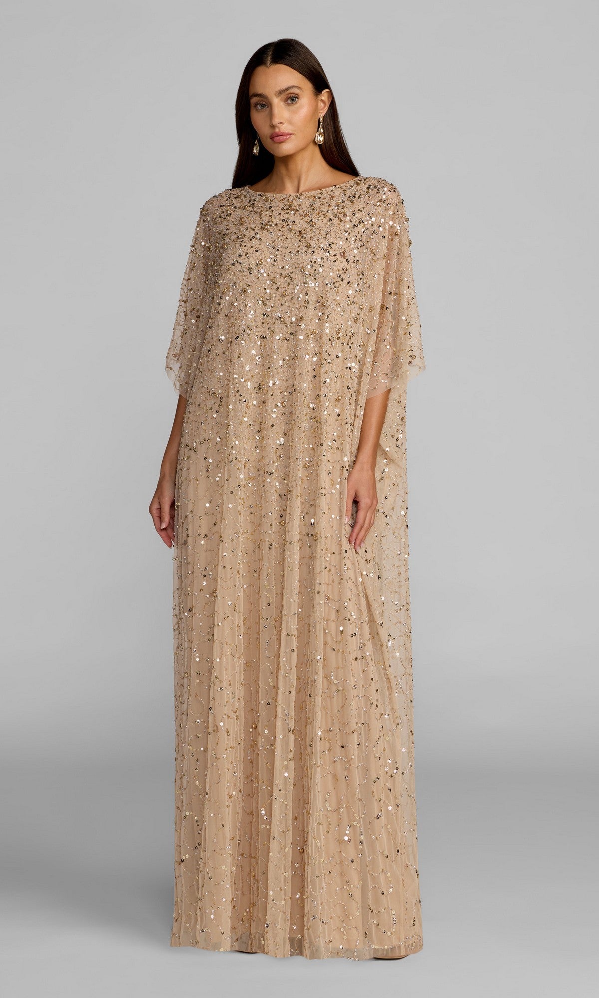 A woman wearing the Mac Duggal 9475 long formal dress in gold, featuring a loose, floor-length silhouette with sequin embellishments, stands against a plain gray background.
