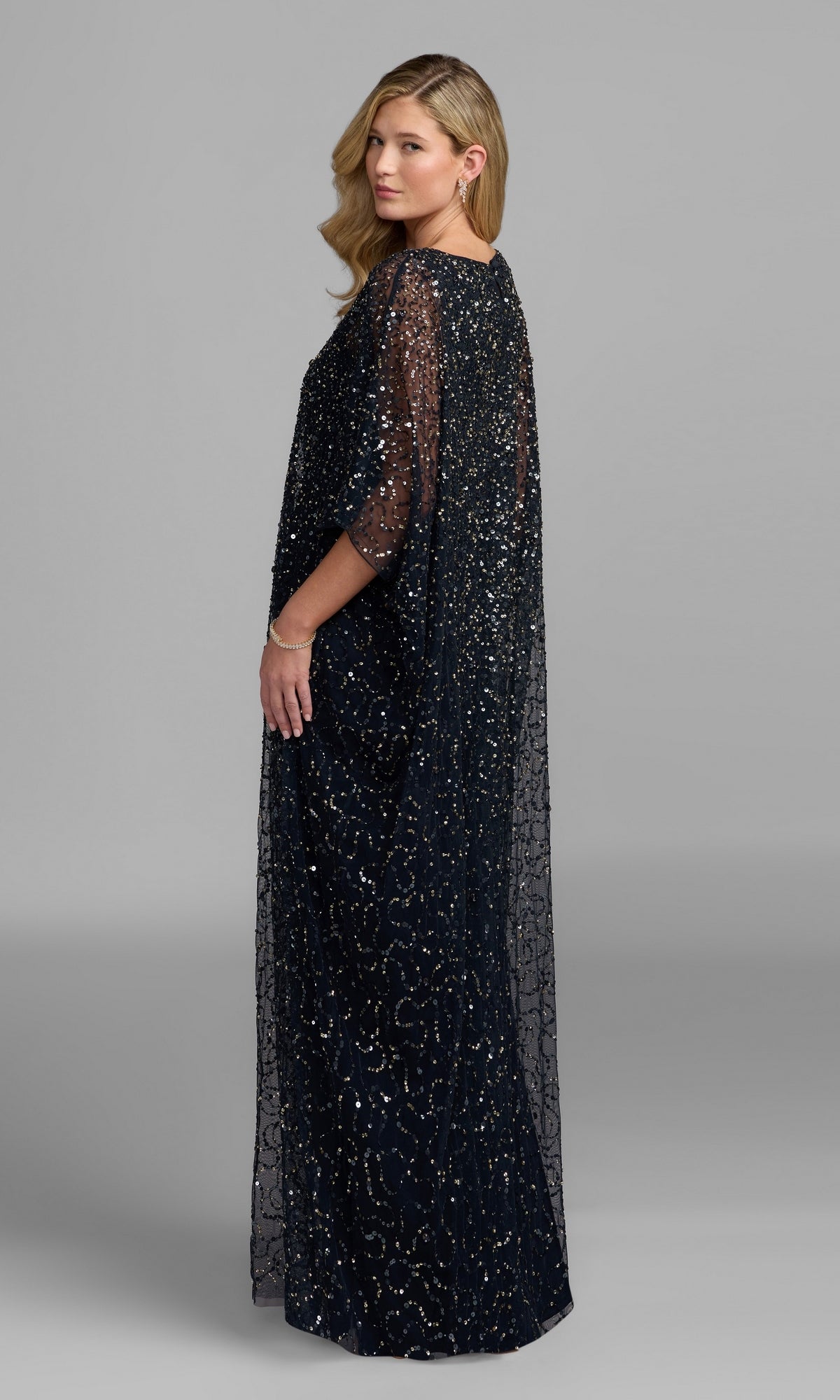 Standing slightly sideways against a plain gray background, the woman wears the Mac Duggal 9475 long formal dress, a floor-length gown adorned with sequin beads and finished with sheer sleeves.
