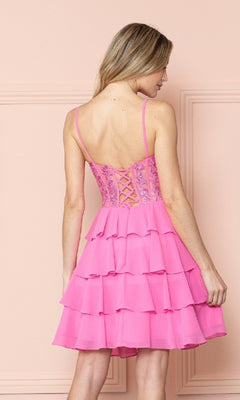 A woman stands with her back to the camera against a light pink wall, wearing the Poly USA 9494 Short Ruffled Lace-Up Hoco Dress—a sleeveless pink homecoming dress featuring a corset-style bodice and lace-up back.