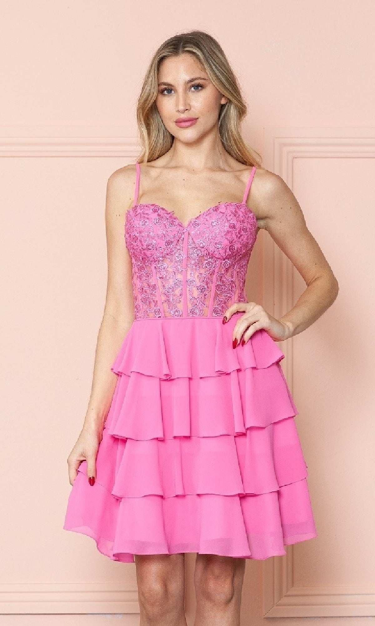 Woman wearing the Short Ruffled Lace-Up Hoco Dress: Poly USA 9494—a sleeveless pink dress with a corset-style bodice and tiered ruffle skirt—stands against a light pink wall.