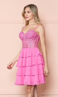 A woman stands against a pale pink wall wearing the Short Ruffled Lace-Up Hoco Dress: Poly USA 9494, featuring a pink corset-style, floral-embellished bodice and layered skirt.
