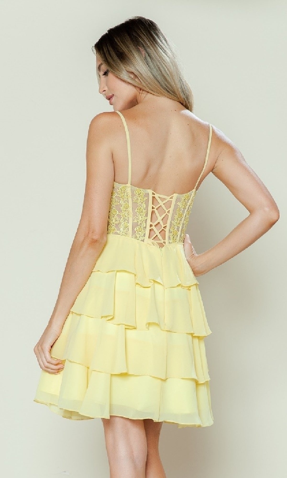 A woman stands with her back to the camera in the Short Ruffled Lace-Up Hoco Dress: Poly USA 9494, a sleeveless, light yellow homecoming dress featuring a corset-style bodice and lace detailing.
