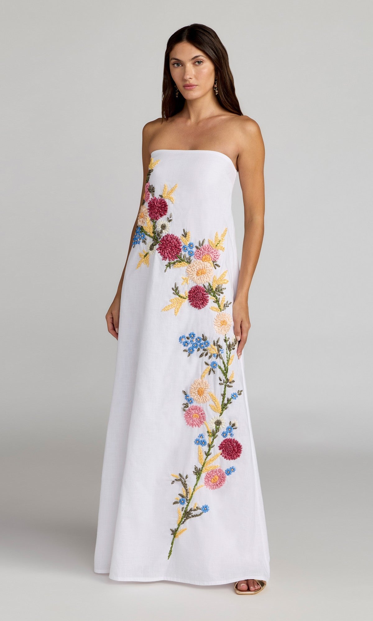 A woman wears the Mac Duggal 9498 Long Strapless Linen Dress in White Multi, featuring colorful floral embroidery, standing against a plain light background.