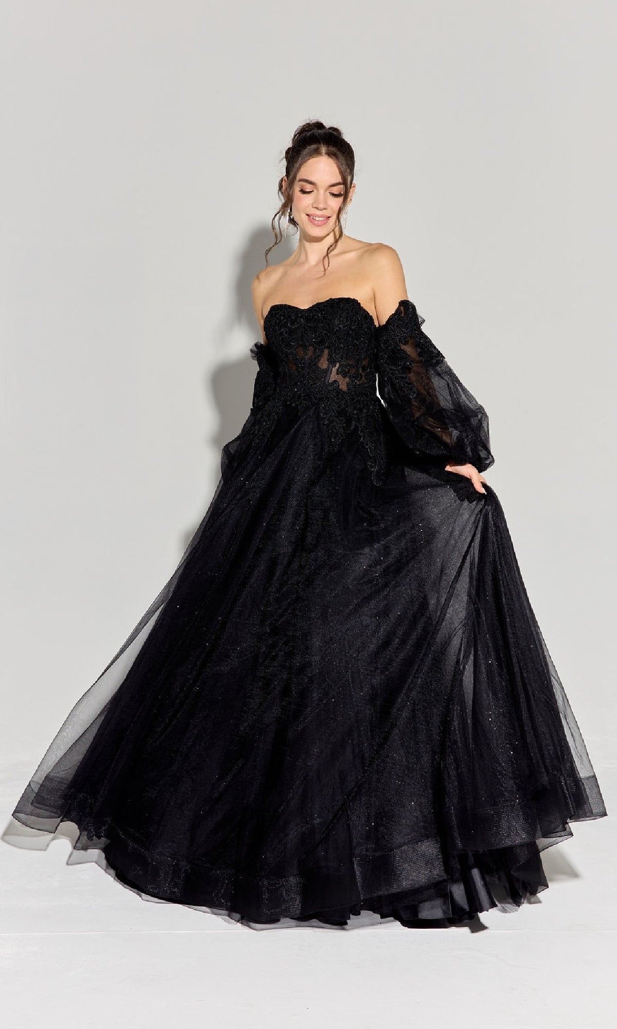 A woman wears the Eureka 9505G Long Sleeve Strapless Prom Ball Gown, featuring sheer off-shoulder sleeves and black embroidery, standing against a plain light background.