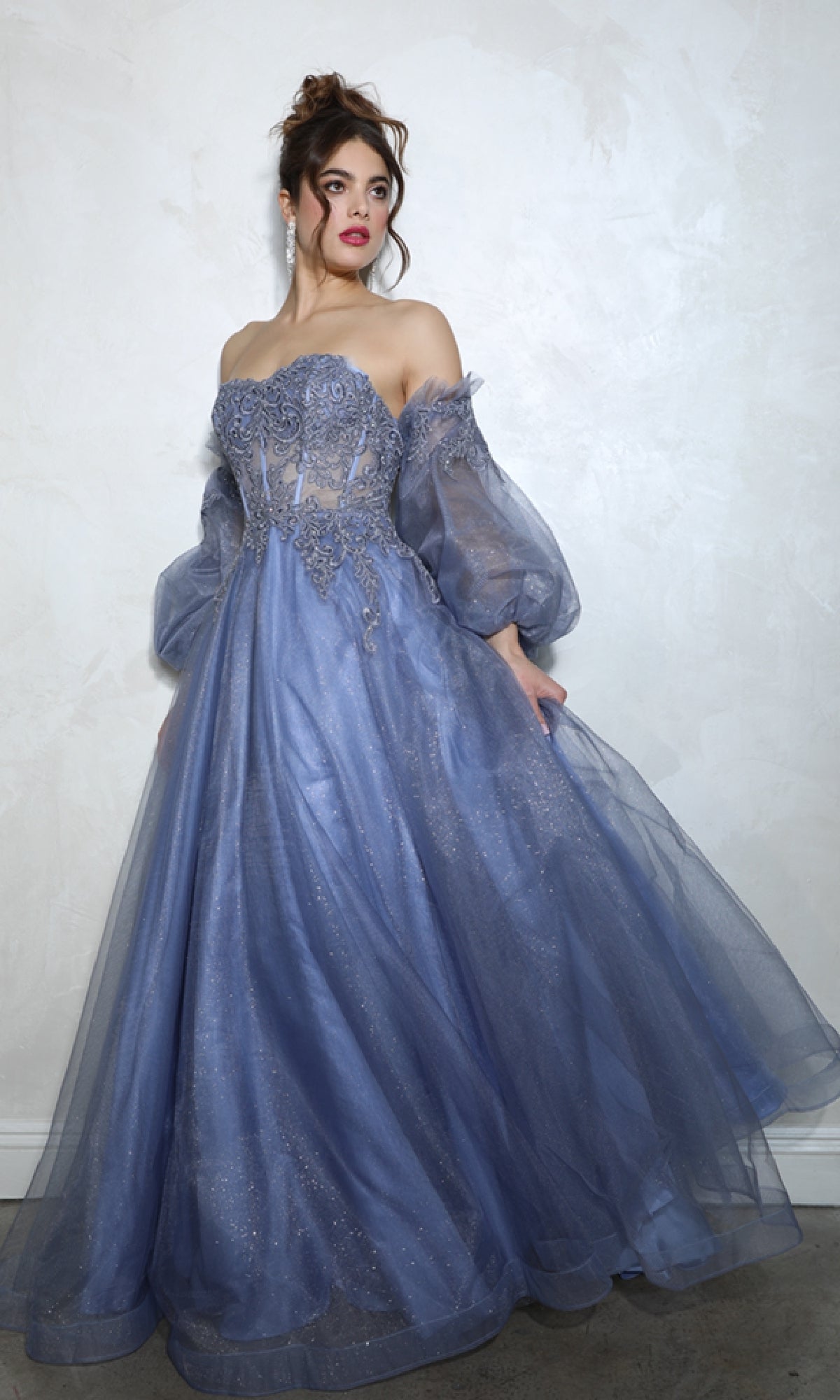 Someone stands against a light wall wearing the Eureka 9505G, a strapless, floor-length prom ball gown with long sheer puffy sleeves and intricate embellishments in lavender.