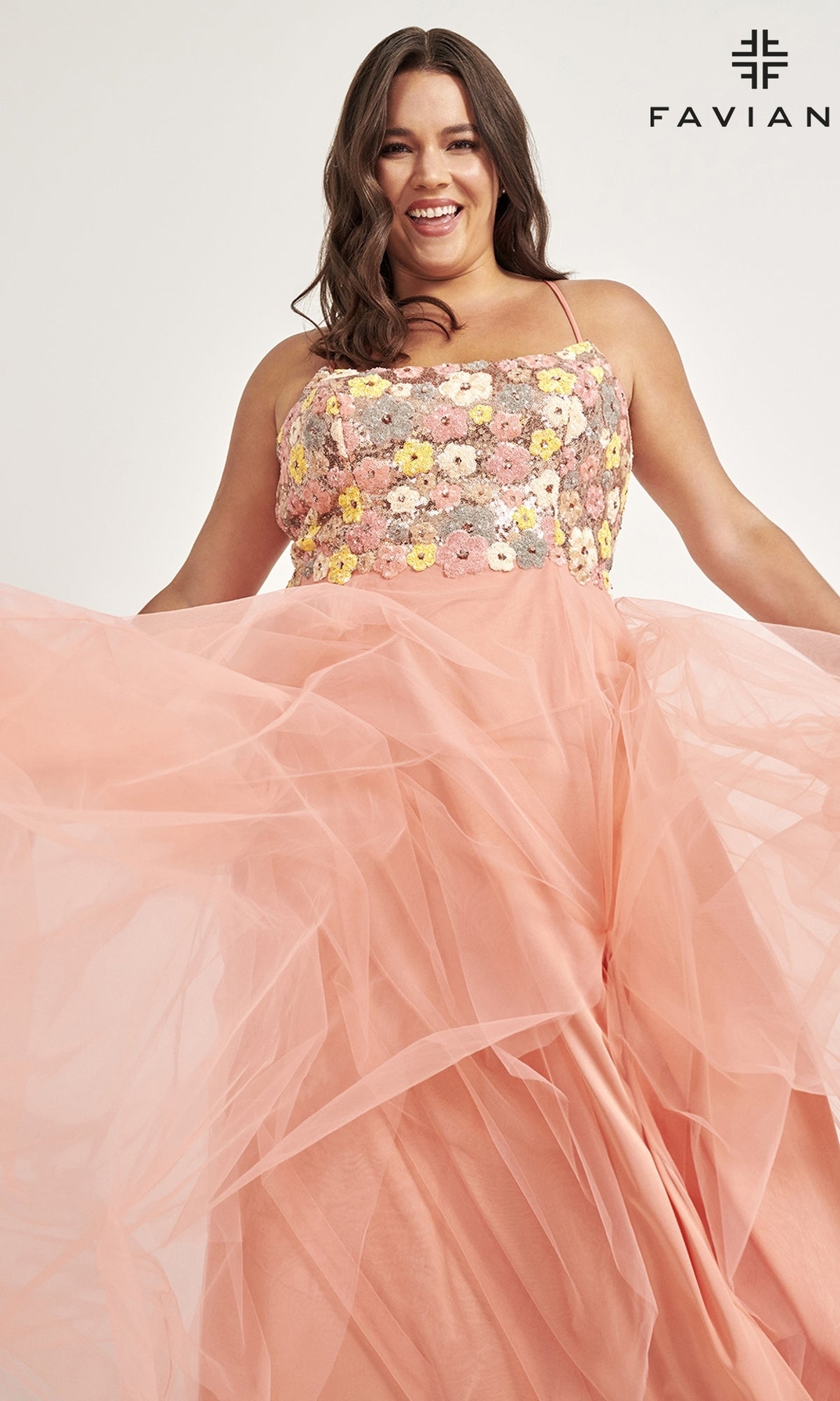 A smiling woman in the Faviana Floral Plus-Size Long Prom Dress 9557 poses, holding out the peach tulle skirt with a floral embroidered bodice. The background is plain with 