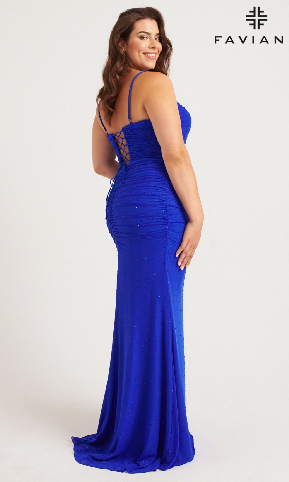 A woman wears the Faviana 9567 Lace-Up Plus-Size Long Prom Dress in royal blue, featuring thin straps, back lacing, subtle embellishments, and a graceful floor-length skirt as she stands sideways and smiles.