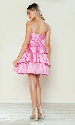 A woman stands on a wooden floor, facing away, wearing the Poly USA 9590 short homecoming dress with a statement bow and pink ruffles, paired with sparkly high-heeled sandals.
