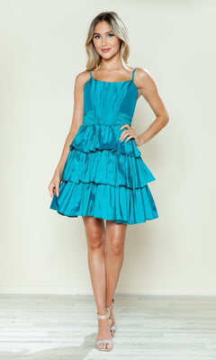 A woman stands indoors against a plain light background, wearing the Poly USA 9590 short homecoming dress with a sleeveless fit-and-flare design, teal tiered ruffle skirt, and nude high-heeled sandals.