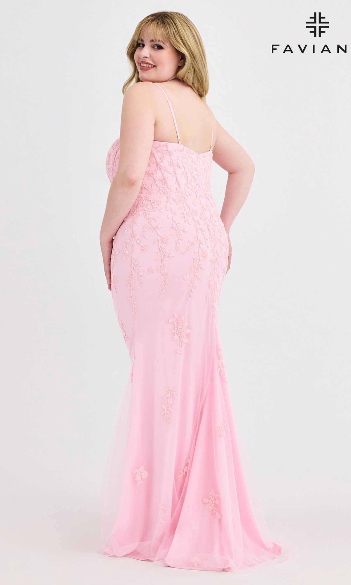 A woman in the Faviana 9604 Plus-Size Beaded-Lace Prom Gown, a light pink, floor-length dress with thin straps, stands facing backward and looks over her shoulder. The plain white backdrop displays the Faviana logo in the top right corner.