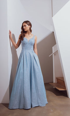 A woman stands indoors wearing the Eureka 9606 Long Prom Dress in ice blue, featuring a sleeveless embroidered bodice and a full, shimmering A-line skirt that gracefully reaches the floor.
