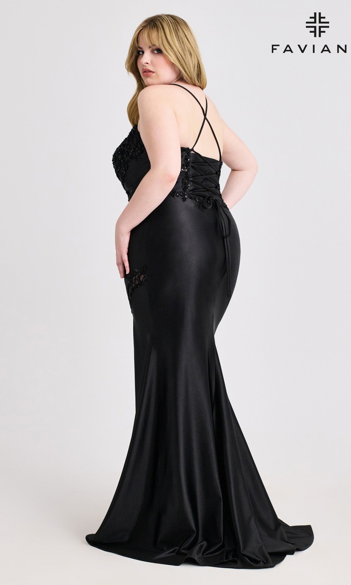 A woman in the Faviana 9606 Lace-Up Plus-Size Long Prom Dress stands facing away, looking over her shoulder. The fitted black gown features a satin skirt, thin cross-back straps, lace-up back detailing, and a slight train.