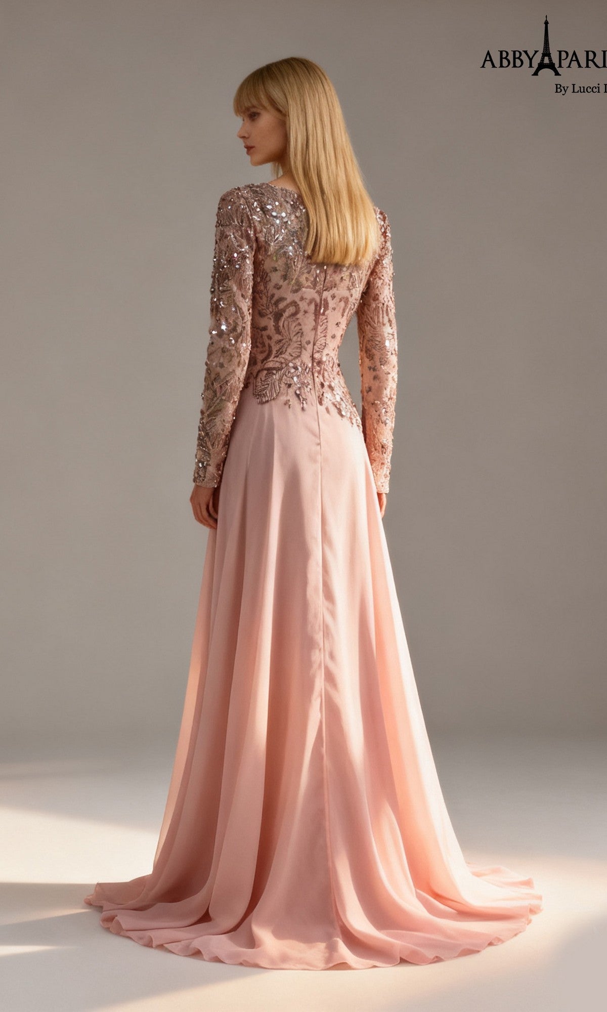 A woman wearing the Long Formal Dress: Abby Paris 96095, featuring a long, light pink A-line silhouette with sequin-embellished sleeves and bodice, stands facing away against a plain background.