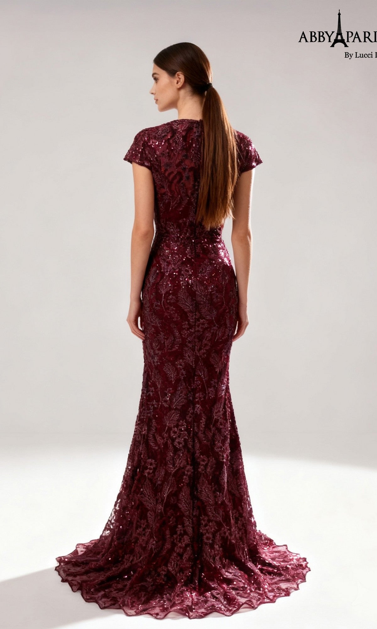 A woman stands facing away, her long straight hair cascading down her back, wearing the Abby Paris 96096—an elegant, floor-length formal dress in burgundy lace with short sleeves and a flattering sheath silhouette—making it an ideal choice for the mother of the bride.