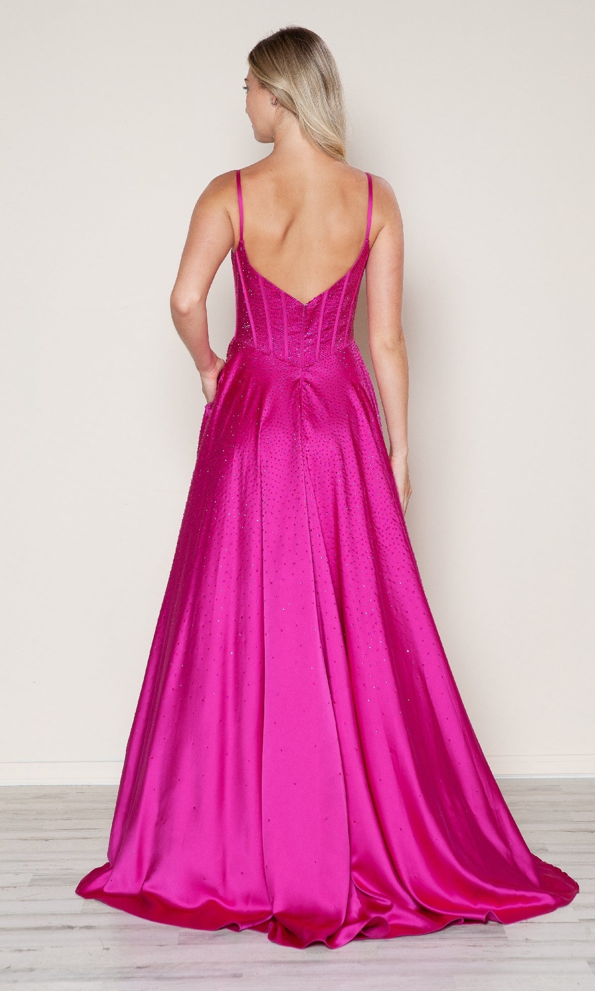 A woman stands facing away, wearing the Poly USA 9610 long prom dress—a floor-length, bright pink satin gown with thin straps, a deep V-shaped back, and an elegant corset bodice.
