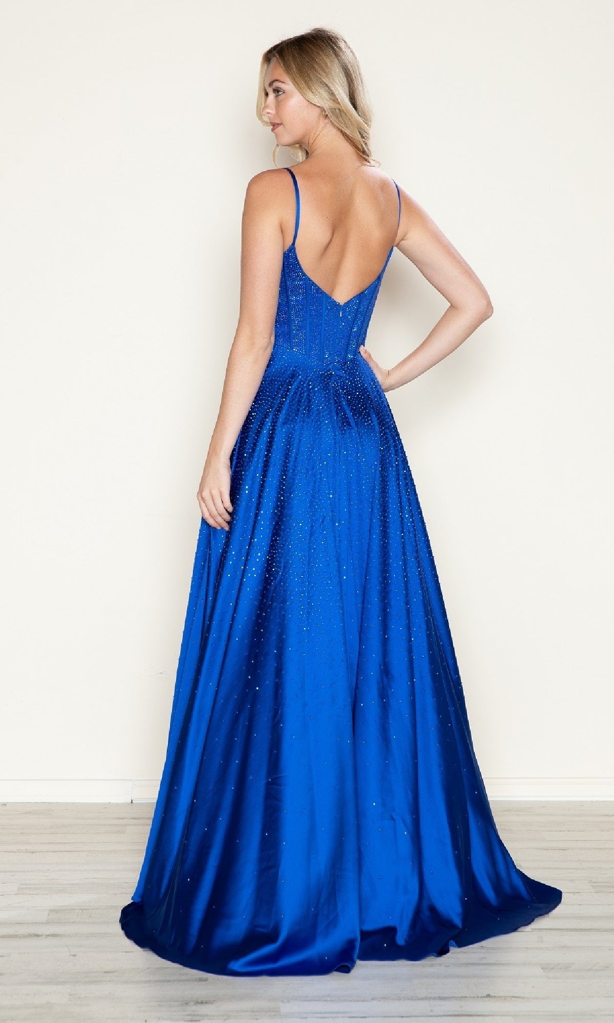 A woman stands indoors facing away from the camera, wearing the Poly USA 9610 long prom dress—a royal blue, floor-length satin A-line gown with thin straps and a low back.