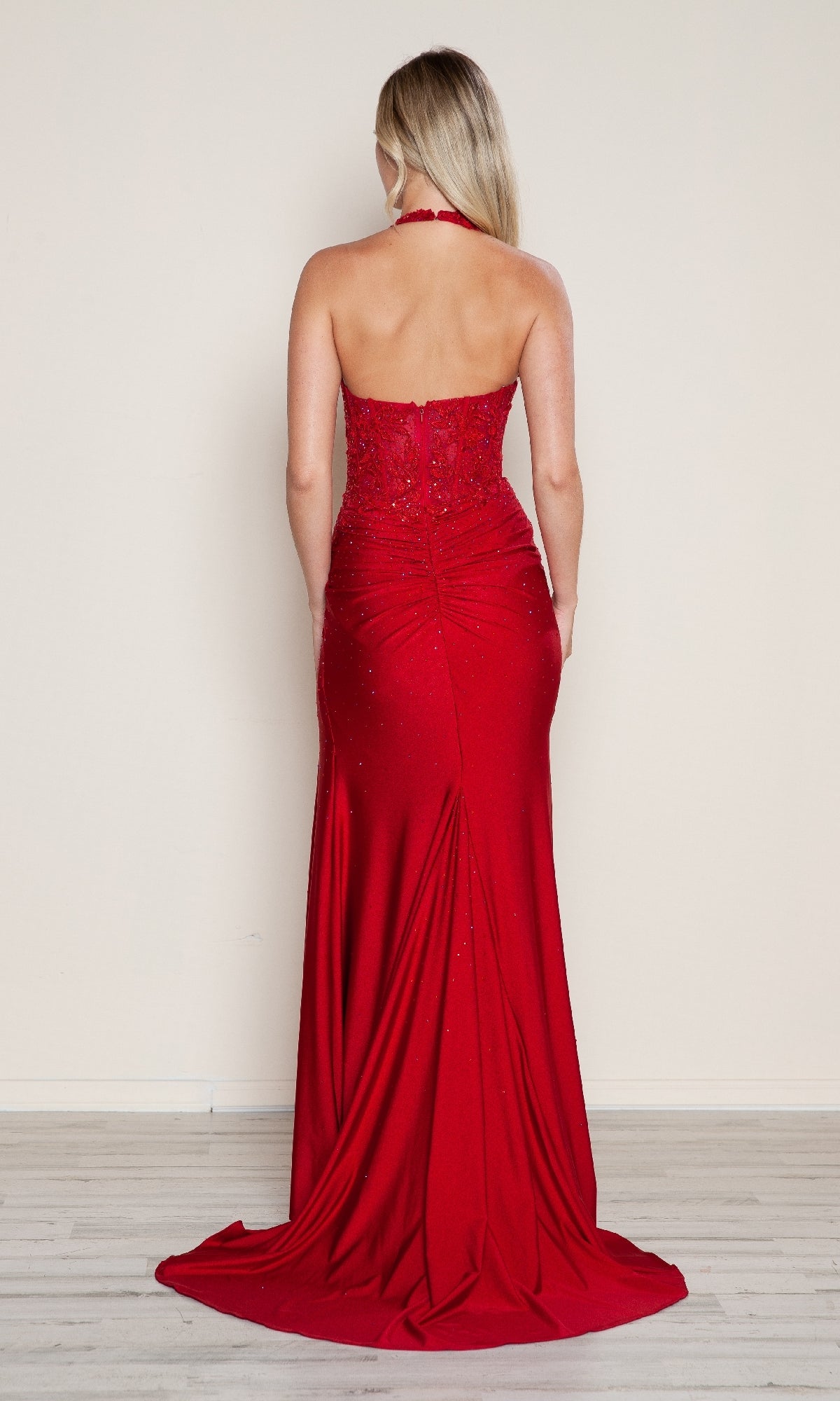 A woman with long blonde hair models the Poly USA 9614 long prom dress—floor-length, strapless, vibrant red with a high leg slit—as she faces away on a light wood floor against a plain beige wall.