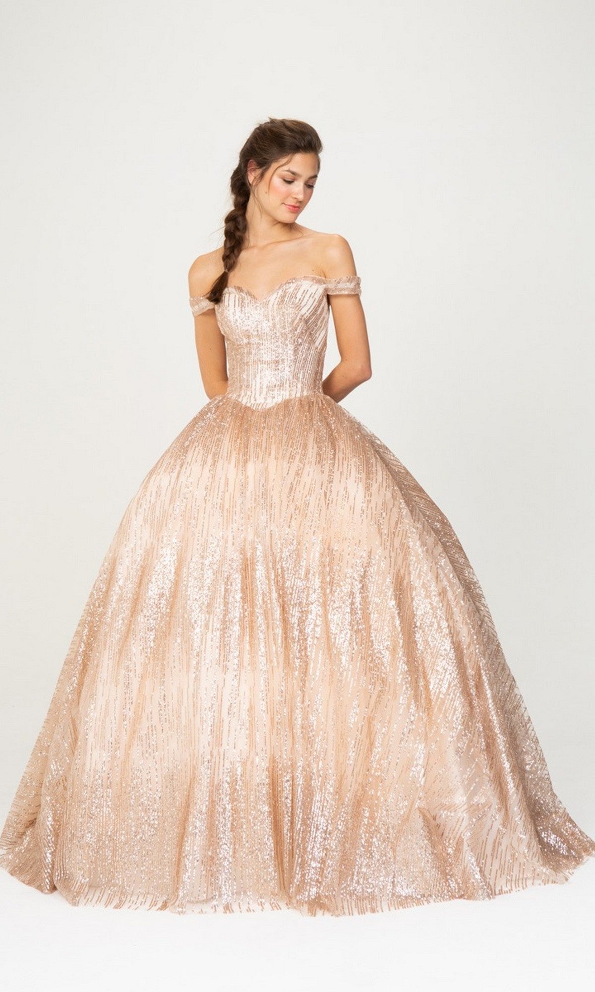 A woman wearing the Quinceanera Dress 9616 By Eureka in Rose Gold, featuring a shimmery, off-the-shoulder fitted bodice and a full ball gown skirt, stands against a plain light background.