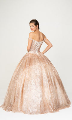 A woman stands against a plain background, looking over her shoulder in the Quinceanera Dress 9616 By Eureka—an off-the-shoulder, light pink ball gown featuring a corset back.