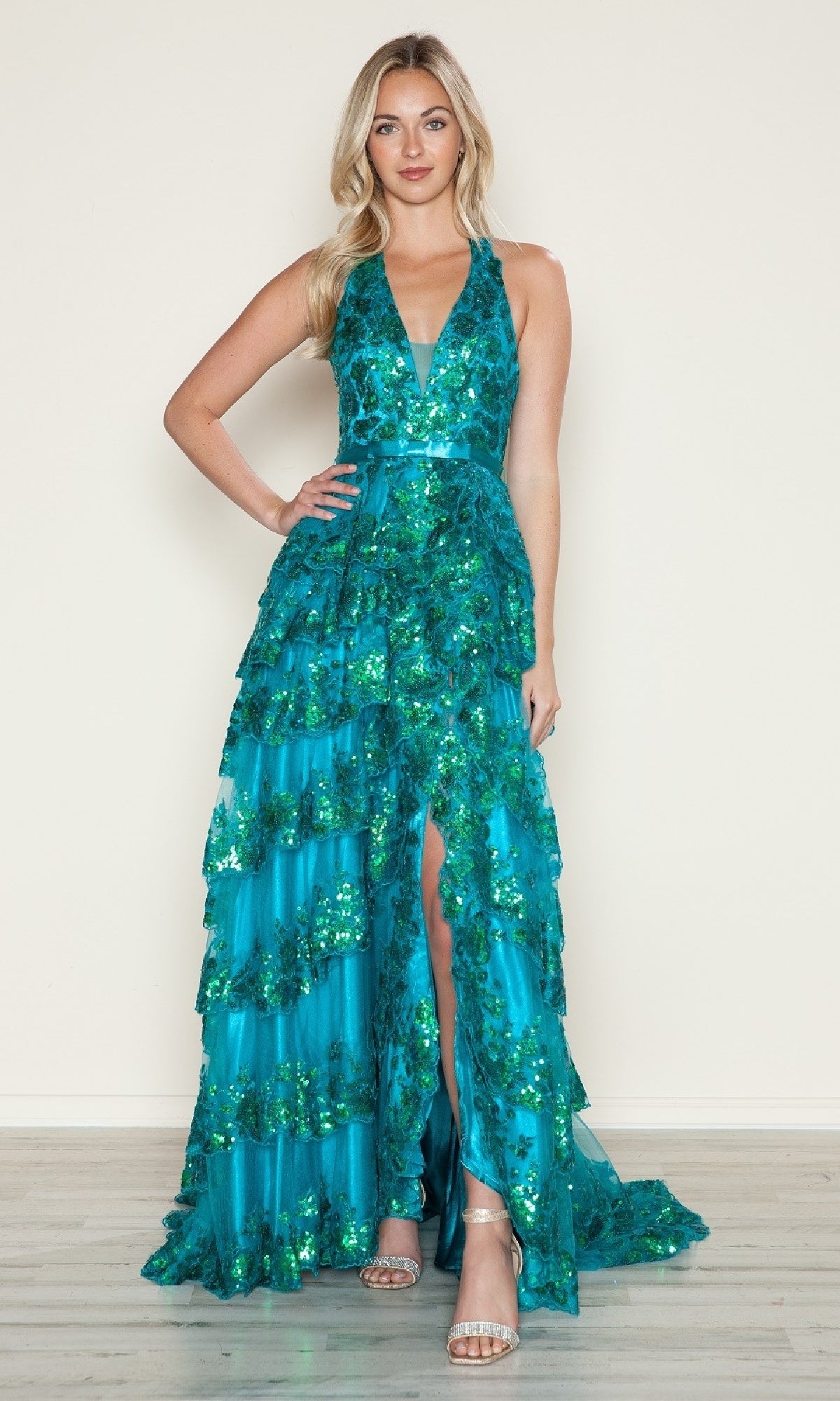 A woman wears the Long Prom Dress: Poly USA 9618, featuring a sleeveless deep V-neck, sequin design, ruffled layers, and high slit, paired with silver heels against a plain background.