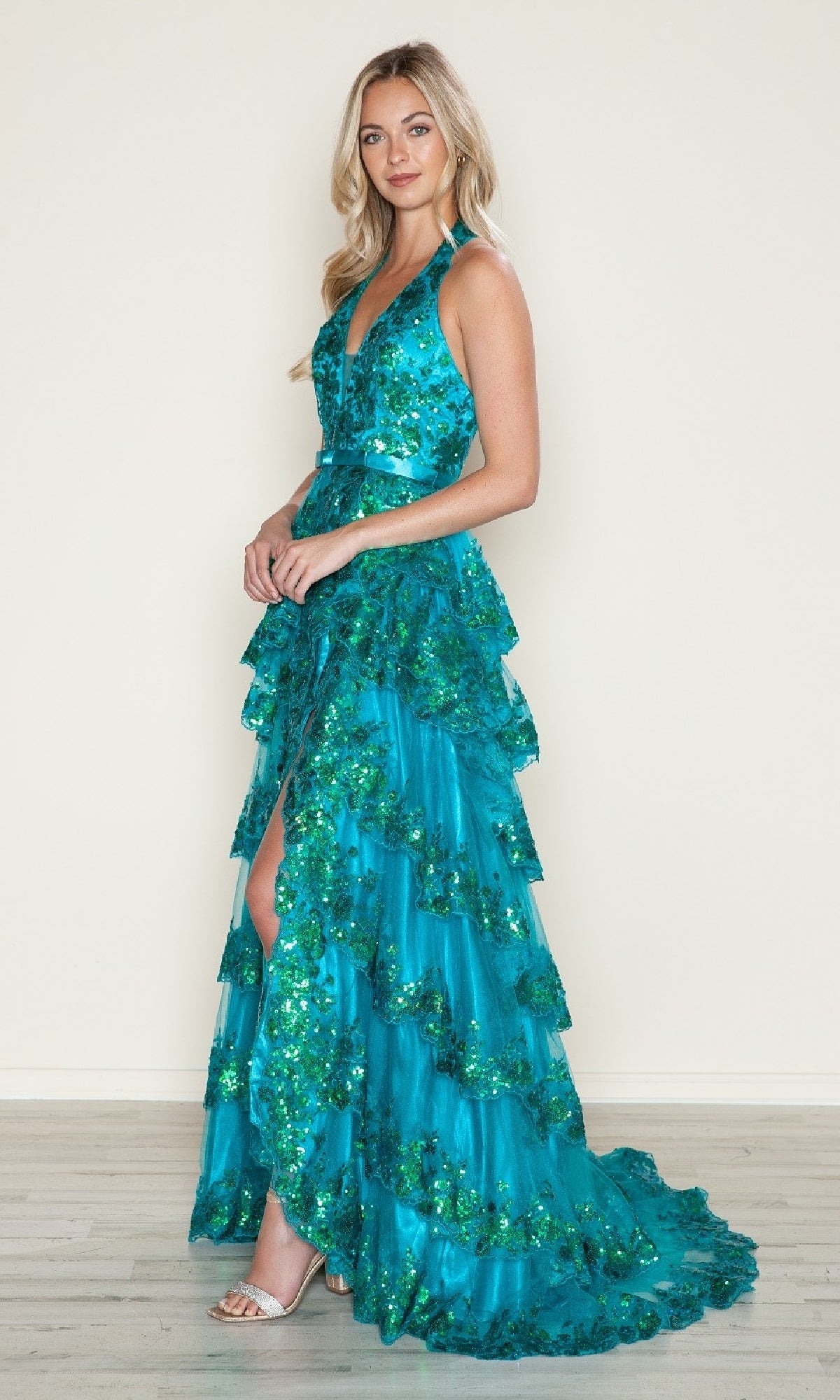 A woman wearing the Poly USA 9618 long prom dress—sleeveless, tiered, turquoise A-line with green sequin accents—stands on a wooden floor against a plain beige wall.