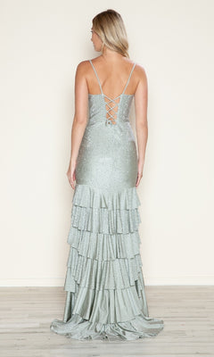 A woman stands on a wooden floor in the Poly USA 9620 Strappy Long Ruffled Prom Dress, featuring thin straps, a lace-up open back, shimmering sequins, and a layered ruffled floor-length skirt against a plain wall.