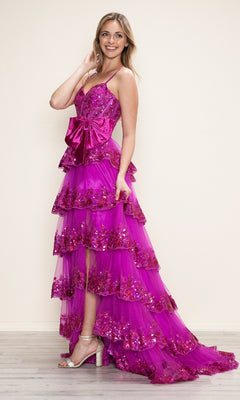 A woman stands indoors wearing the Long Prom Dress: Poly USA 9626, a bright pink A-line gown with a sequin corset bodice, tiered skirt, large satin waist bow, and silver high-heeled sandals.