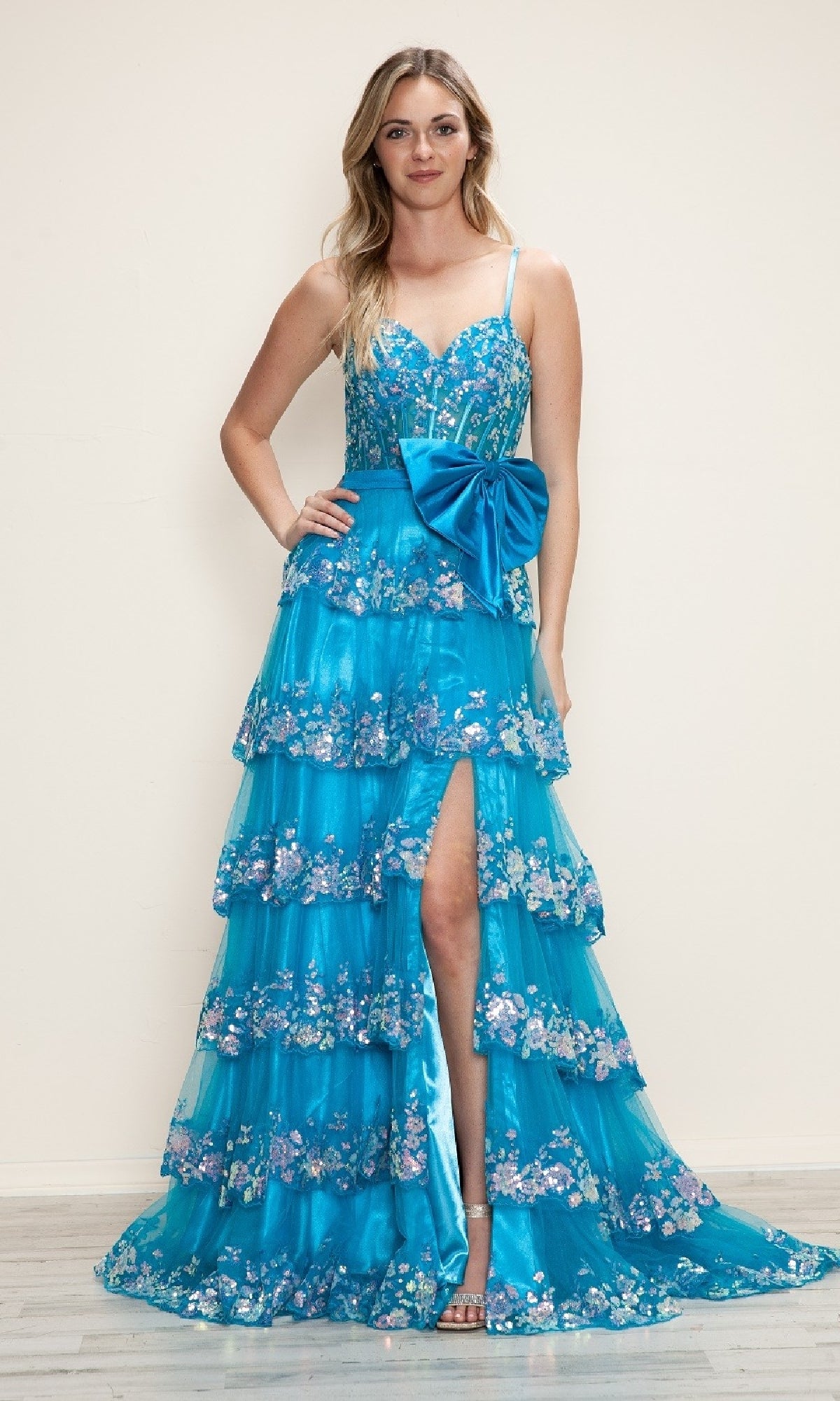 A woman wears the Poly USA 9626 long prom dress, featuring an A-line silhouette, tiered blue skirt with floral details, a large waist bow, and thigh-high slit, standing against a plain light background.