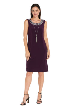 Model wears the R&M Richards 9626P Petite Two-Piece Short Party Dress—sleeveless purple with decorative neckline—paired with black heels and a long pendant necklace, posed against a white background.