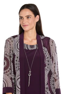 A woman with straight dark hair, facing slightly to the side and smiling, wears the R&M Richards 9626P Petite Two-Piece Short Party Dress in eggplant, paired with a long purple scarf and a silver pendant necklace.