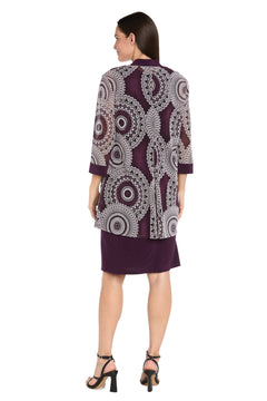 A woman faces away, modeling the Petite Two-Piece Short Party Dress: R&M Richards 9626P—a short purple dress with a patterned, long-sleeve overlay and black heeled sandals, set against a white background.