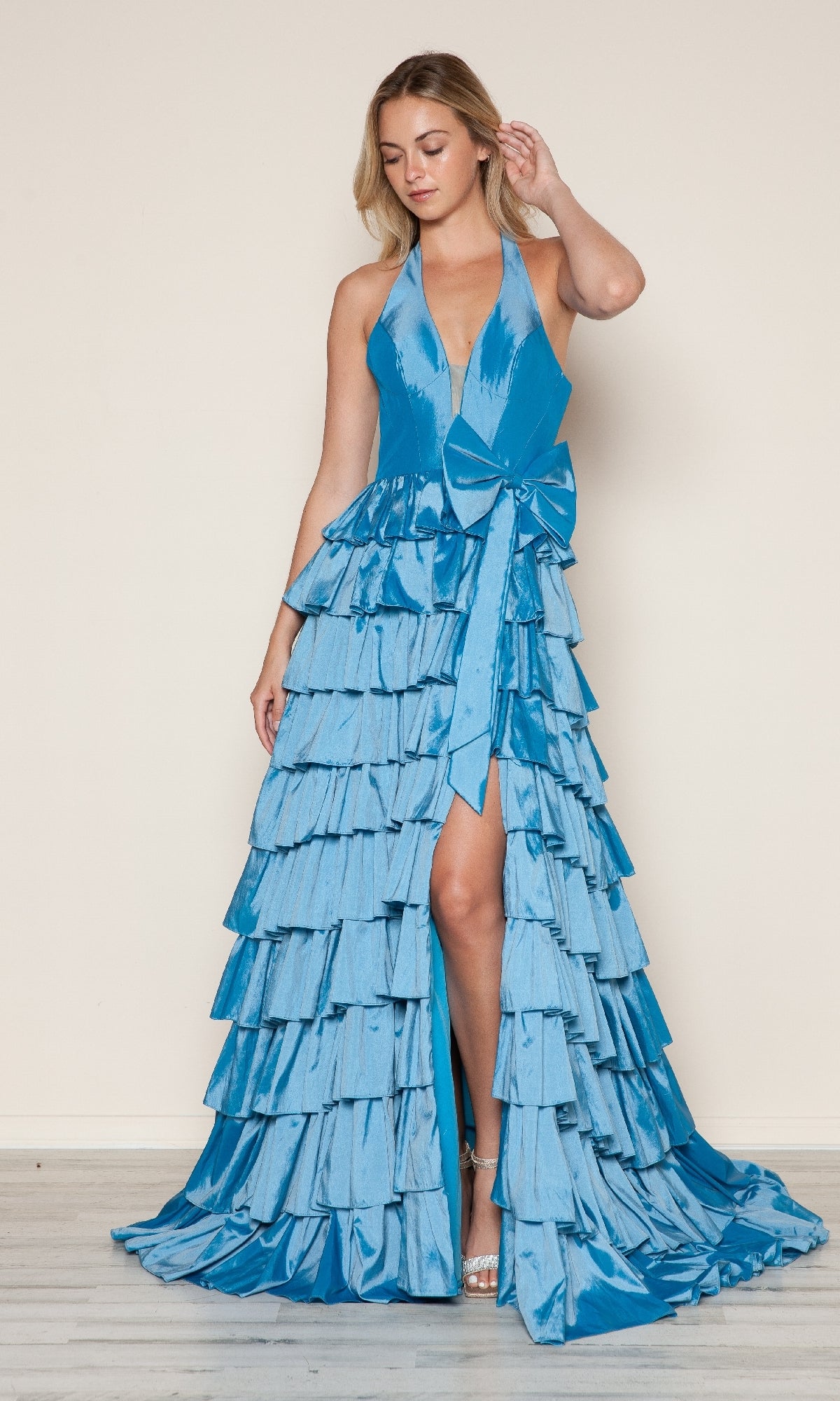A woman poses in a shiny blue Ruffled Long Halter Prom Dress (Poly USA 9642) featuring a deep V-neck, large waist bow, and multiple ruffled layers, set against a plain light background.