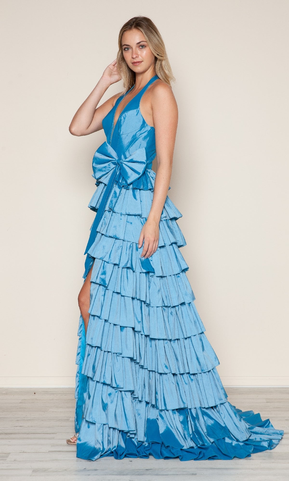 A woman models the Poly USA 9642 Ruffled Long Halter Prom Dress, featuring a floor-length tiered blue design with a large front bow, against a plain light background.