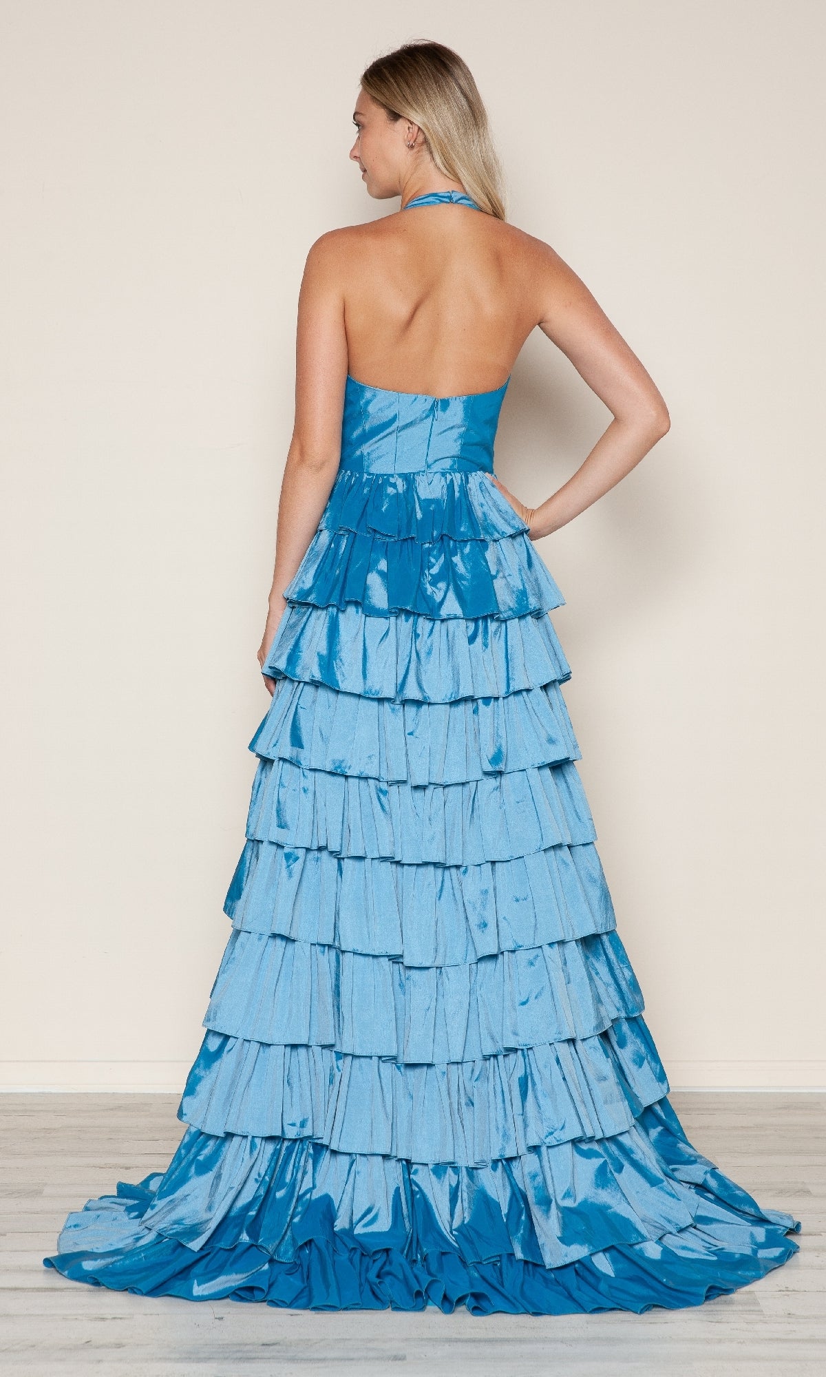 A woman stands with her back to the camera in the Poly USA 9642 Ruffled Long Halter Prom Dress, featuring blue tiered ruffles and a dramatic floor-length train.