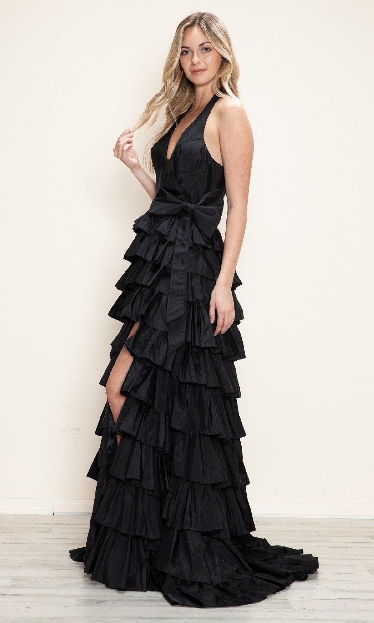 A woman poses against a light wall on a wooden floor, wearing the Ruffled Long Halter Prom Dress: Poly USA 9642, featuring a black tiered ruffle design and a bow at the waist.