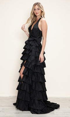 A woman poses against a light wall on a wooden floor, wearing the Ruffled Long Halter Prom Dress: Poly USA 9642, featuring a black tiered ruffle design and a bow at the waist.