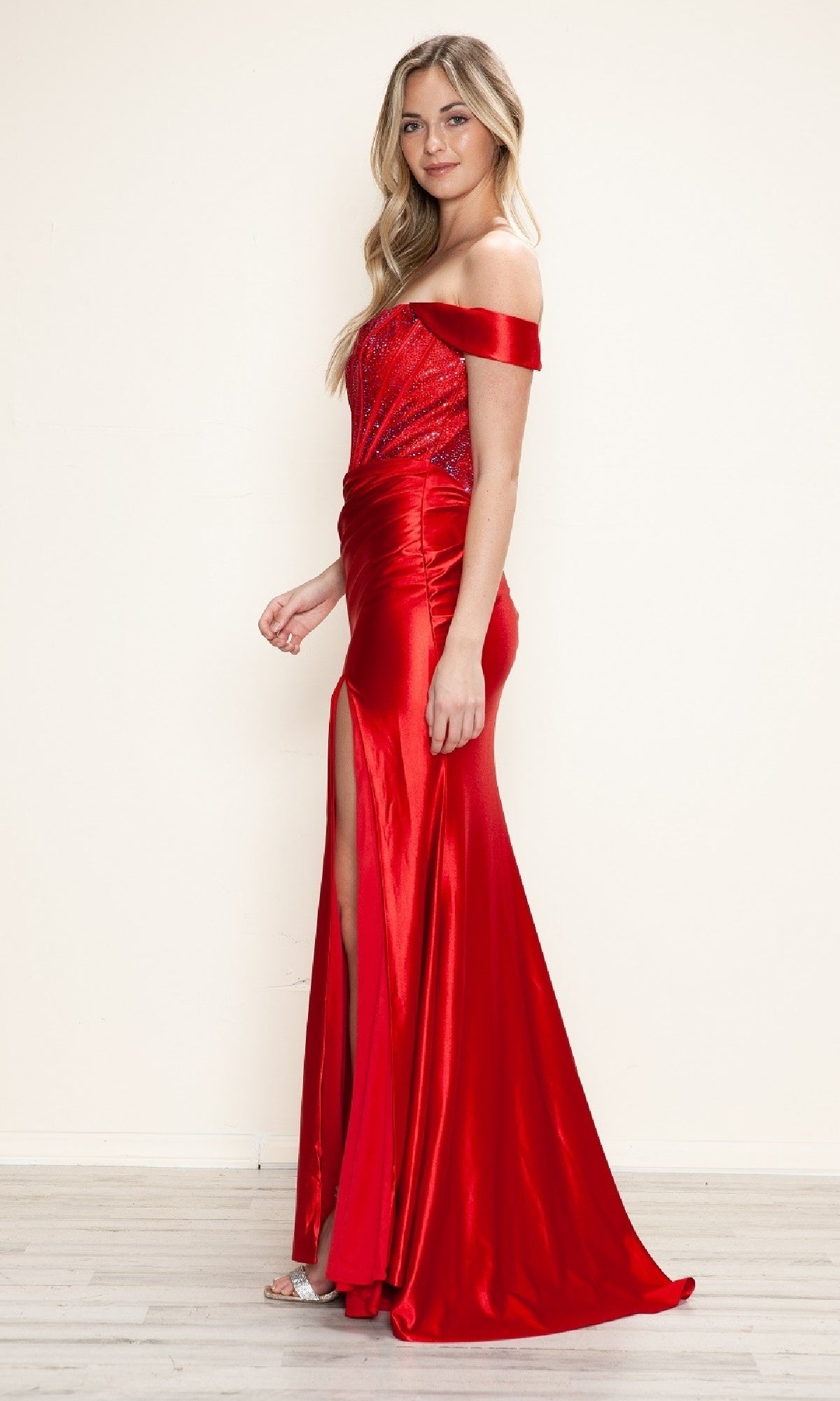A woman in the Off-Shoulder Long Corset Prom Dress: Poly USA 9656—shiny red, with a beaded corset and thigh-high slit—stands on a light wooden floor against a plain cream wall.