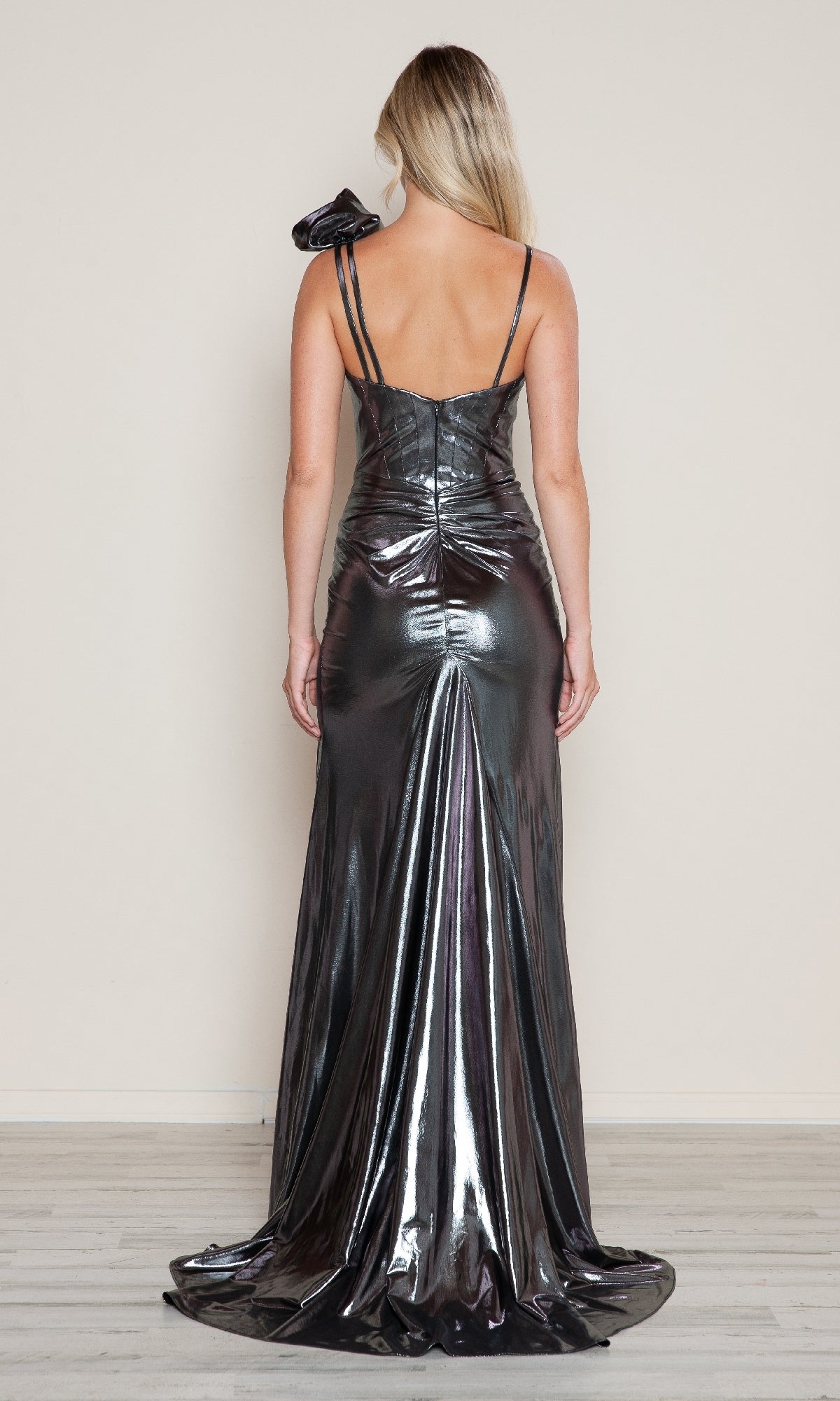 A woman faces away, wearing the Poly USA 9680 Ruffle Shoulder Metallic Prom Gown—a shiny silver evening dress with thin straps and a ruffled bow detail on one shoulder.