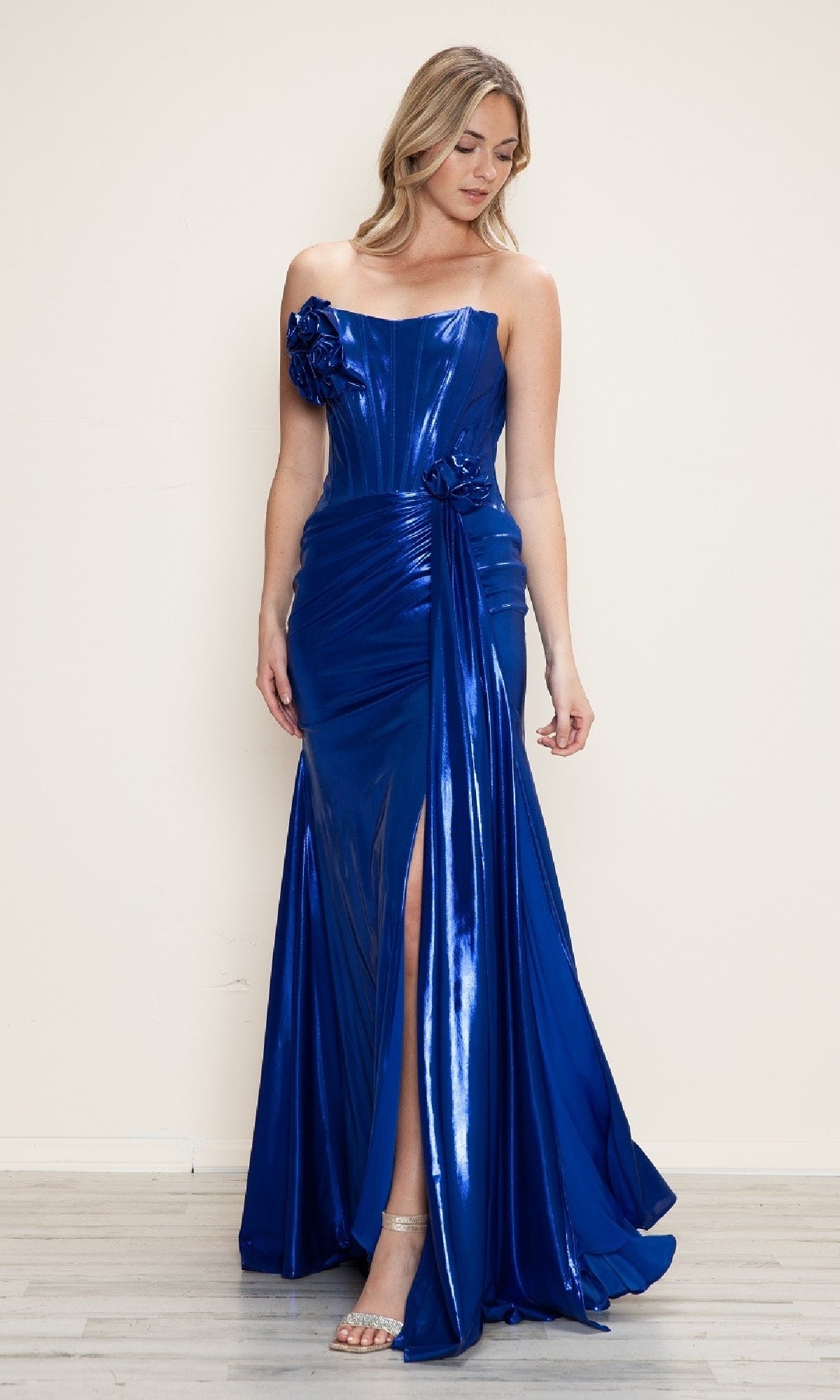 A woman in the Strapless Long Metallic Prom Dress: Poly USA 9682, featuring a shiny blue finish, floral accents, thigh-high slit, and paired with silver heels, stands against a plain light-colored wall.