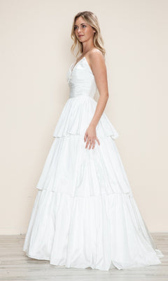 A woman stands indoors wearing the Long Prom Dress: Poly USA 9694, a white, floor-length gown with a fitted bodice and tiered a-line skirt.