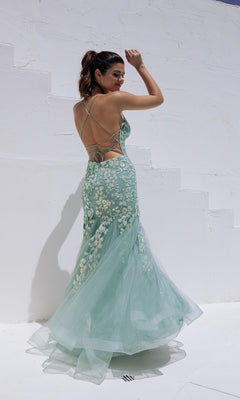 A woman in the Sequin-Floral Long Mermaid Prom Dress: Eureka 9696 poses outdoors against a white textured wall, her hair tied back and one arm raised.
